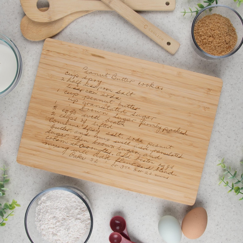 Large rectangular bamboo board shown with a "Peanut Butter Cookies" recipe engraved on it. A few kitchen items such as flour, milk, brown sugar, measuring tools, & spoons are displayed around the cutting board