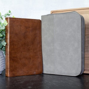May include: Two leather-bound journals, one brown and one gray, are displayed on a black surface. The brown journal has a spine with the words "HOLY BIBLE" embossed on it. The gray journal has a zipper closure.