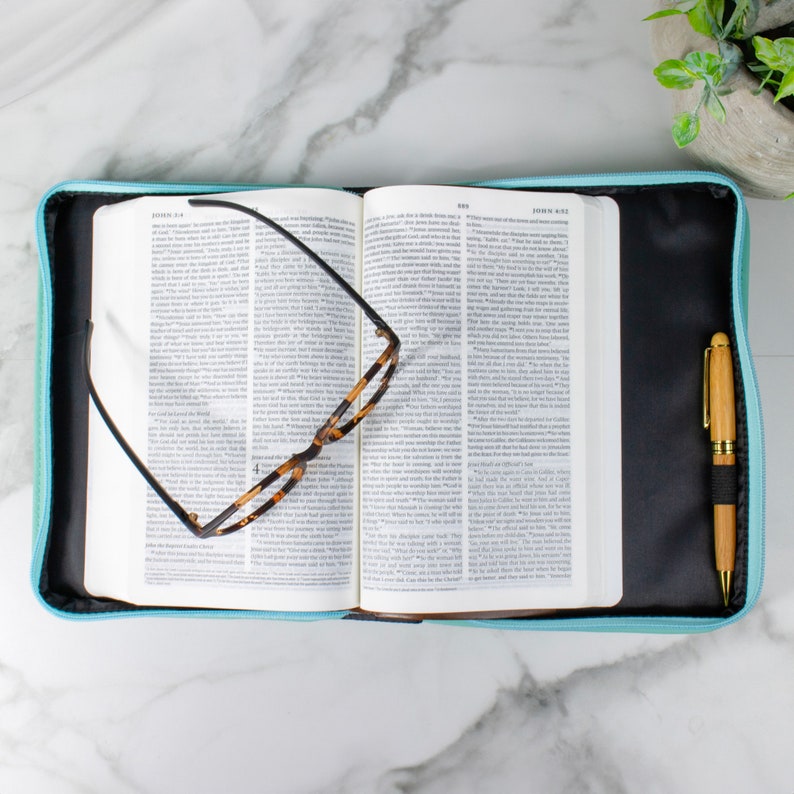 May include: A light blue zippered Bible cover with a black interior. A pair of tortoiseshell glasses and a wooden pen are resting on the open Bible inside the cover.
