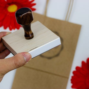 A person’s hand is holding up a wood handle stamp in the air. In the background there is a brown paper bag with a logo stamp impression and two red flowers laying on both sides of the brown paper bag.