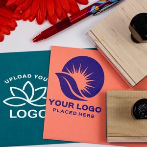 One large wood handle stamp and another smaller wood handle stamp sit next to each other on top of two sheets of paper. Two sample logos are used as an impression on the sheets of paper in white and blue ink. A red pen and flower sit above the paper.