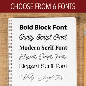 May include: A spiral-bound notebook displays font options. The text "CHOOSE FROM 6 FONTS" is at the top. Below, six font styles are shown: Bold Block, Curly Script, Modern Serif, Elegant Script, Elegant Serif, and Vintage Script. A pencil rests on the right.