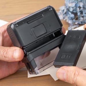 May include: A black self-inking stamp with a clear base, shown open with the ink cartridge removed. The stamp is held by hands, with a piece of paper and blue flowers in the background.