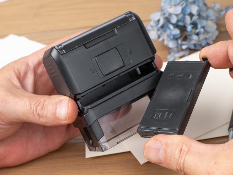 May include: A black self-inking stamp with a clear base, shown open with the ink cartridge removed. The stamp is held by hands, with a piece of paper and blue flowers in the background.