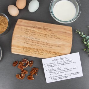 Small maple cutting board is shown engraved with a  "Glazed Pecans" recipe on it with the original handwritten recipe card next to it. Some kitchen items such as eggs, milk, brown sugar, & pecans are displayed around the cutting board and recipe card