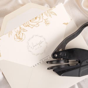 May include: An ivory invitation with gold floral accents and the name "Washington" is partially inside an ivory envelope. A black Trodat embosser sits on the invitation. The date "06.17.23" is also visible.