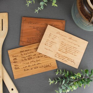 A dark, medium, & light wood grain recipe card is displayed with a different recipe engraved onto each card. Some wooden kitchen spoons & decorative leaves are displayed around the wooden recipe cards.