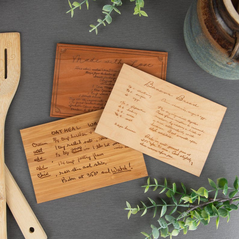 A dark, medium, & light wood grain recipe card is displayed with a different recipe engraved onto each card. Some wooden kitchen spoons & decorative leaves are displayed around the wooden recipe cards.