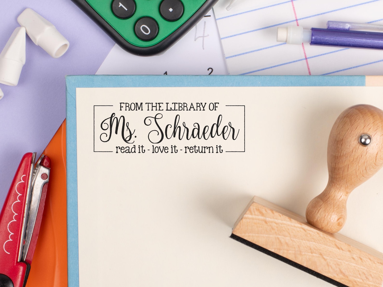 A personalized bookplate stamp reads “From the Library of Ms. Schraeder” in a combination of typewriter-style and elegant script fonts, enclosed within a minimalist box border. Below the name, a smaller line of text says, “read it – love it – return it.” The stamp is applied to a cream-colored page framed in light blue, with a wooden-handled stamp tool resting nearby. The scene includes scattered school supplies: a red stapler, a green calculator, pen parts, erasers, and a sheet of lined paper