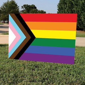 May include: A rectangular yard sign featuring the Progress Pride flag, with a rainbow design and additional stripes representing marginalized groups. The sign is displayed on a grassy lawn with metal stakes.