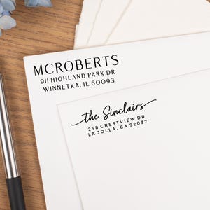 May include: White stationery with printed addresses for McRoberts and the Sinclairs. The McRoberts address is 911 Highland Park Dr, Winnetka, IL 60093. The Sinclairs address is 258 Crestview Dr, La Jolla, CA 92037. A silver pen rests on a wooden surface.