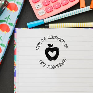 May include: A white notebook with black lines and a black rubber stamp with a red apple and the text "FROM THE CLASSROOM OF MRS. Flenderson".