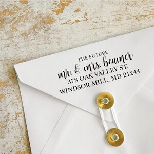 May include: A white envelope with the text "THE FUTURE mr. & mrs. beamer 378 OAK VALLEY ST. WINDSOR MILL, MD 21244" printed in black. The envelope is secured with two gold-colored fasteners and a white string.