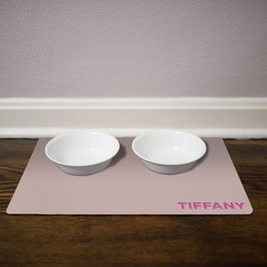May include: A light pink silicone pet food mat with two white bowls and the name "TIFFANY" printed in pink.