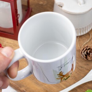 May include: White ceramic mug with a deer illustration. The mug has a handle and a smooth, glossy finish. The deer is depicted in shades of brown, green, and white. Other items in the image include a red lantern, a white spoon, and pine cones.