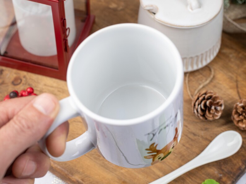 May include: White ceramic mug with a deer illustration. The mug has a handle and a smooth, glossy finish. The deer is depicted in shades of brown, green, and white. Other items in the image include a red lantern, a white spoon, and pine cones.