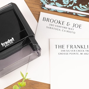 May include: A black Trodat printy 4914 stamp next to two white cards with names and addresses printed in black. A patterned notebook is in the background. The cards read "BROOKE & JOE" and "THE FRANKLINS".