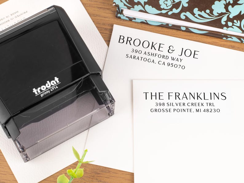 May include: A black Trodat printy 4914 stamp next to two white cards with names and addresses printed in black. A patterned notebook is in the background. The cards read "BROOKE & JOE" and "THE FRANKLINS".