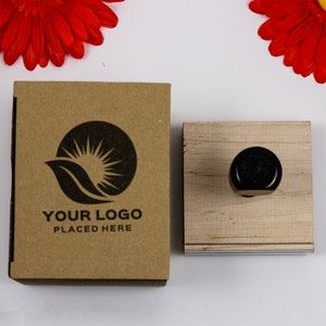 A sample logo is used as an impression on a brown box in black ink. A wood handle stamp is placed next to the brown box. Two red flowers sit above the brown box and the wood handle stamp.