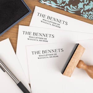 May include: A wooden address stamp with the text "THE BENNETS" and address information printed on white paper. An ink pad and pen are also visible. The paper has the address: 904 CLIFFSIDE DR WAYZATA, MN 55391.