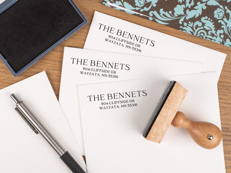 May include: A wooden address stamp with the text "THE BENNETS" and address information printed on white paper. An ink pad and pen are also visible. The paper has the address: 904 CLIFFSIDE DR WAYZATA, MN 55391.