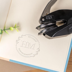 May include: A black and silver book embosser rests on an open book with a light blue border. The embossed design reads "EX LIBRIS" above the initials "HM" within a laurel wreath. The book's pages are cream-colored, and a sprig of greenery is in the upper left.