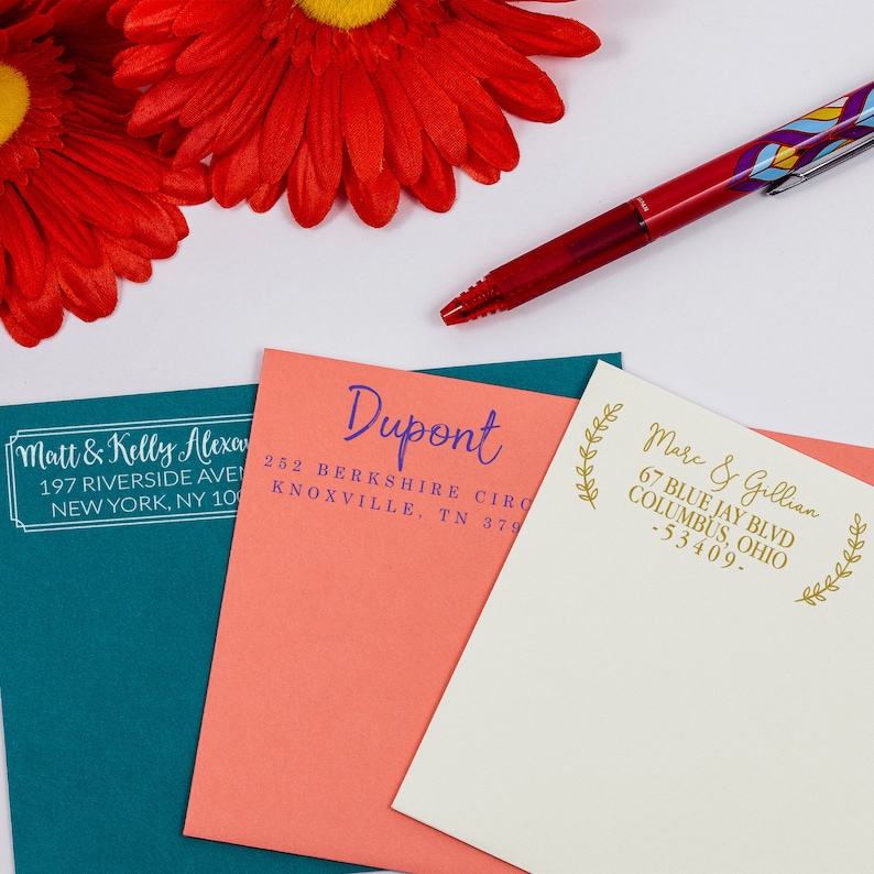 A blue, pink, and white envelope with various personalized address stamp impressions in an assortment of ink colors. Each stamp design includes a custom name and address. A red pen and two flowers are placed above the envelopes.