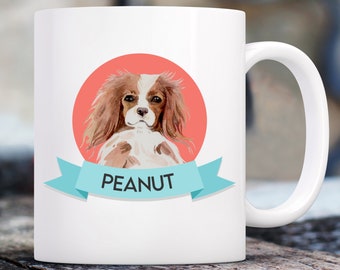 May include: White ceramic mug with a watercolor illustration of a Cavalier King Charles Spaniel dog with brown and white fur. The dog is surrounded by a red circle and a blue ribbon with the name "PEANUT" written on it.