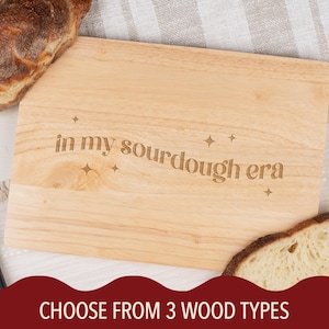 May include: A light-colored wooden cutting board with the phrase "in my sourdough era" engraved on it. The board is rectangular and surrounded by bread and a bread knife. The bottom of the image says "CHOOSE FROM 3 WOOD TYPES".