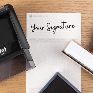 May include: A black Trodat printy 4913 stamp, a wooden handled stamp, and a black ink pad are on a wooden surface. A white document with the words "Your Signature" is also present.