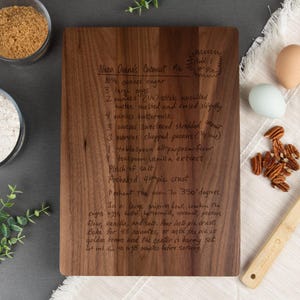 Large walnut cutting board shown with a "Nana Deane's Coconut Pie" recipe engraved on it. Some kitchen items such as eggs, flour, brown sugar, & pecans are displayed around the cutting board with some decorative leaves in the left corner
