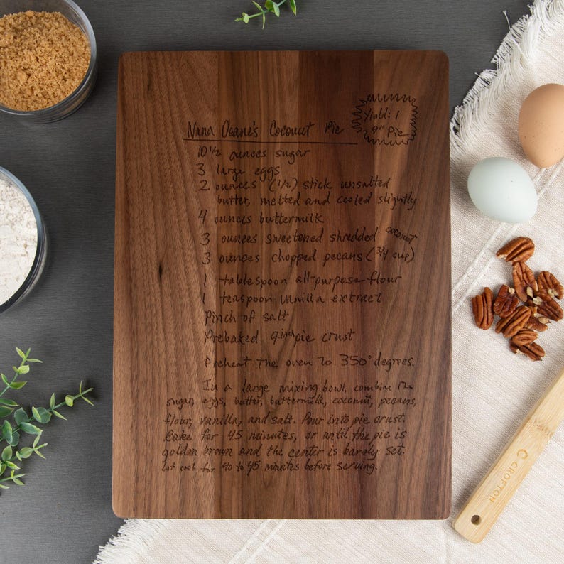 Large walnut cutting board shown with a "Nana Deane's Coconut Pie" recipe engraved on it. Some kitchen items such as eggs, flour, brown sugar, & pecans are displayed around the cutting board with some decorative leaves in the left corner