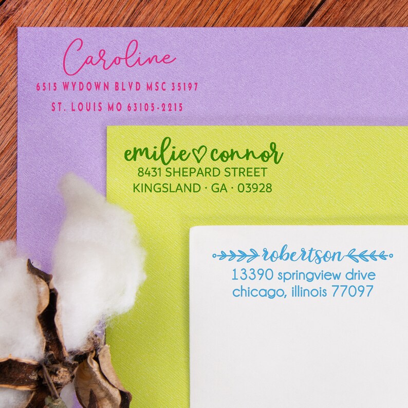A purple, yellow and white envelope fanned out to show each envelope stamped in a different address design all stamped in different ink colors. The purple envelope stamped in pink ink, the yellow stamped in green ink, and the pink stamped in blue ink