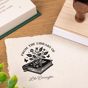 May include: A wooden stamp with a floral book design and the text "FROM THE LIBRARY OF Lila Carrington" is shown on a textured paper. A wooden stamp handle and a book are also visible. The stamp is likely used for personalizing books or stationery.
