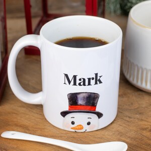 May include: White ceramic mug with the name "Mark" printed in black, featuring a snowman design with a black top hat and orange carrot nose. The mug is filled with dark liquid, likely coffee or tea.