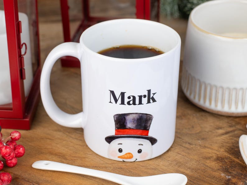 May include: White ceramic mug with the name "Mark" printed in black, featuring a snowman design with a black top hat and orange carrot nose. The mug is filled with dark liquid, likely coffee or tea.