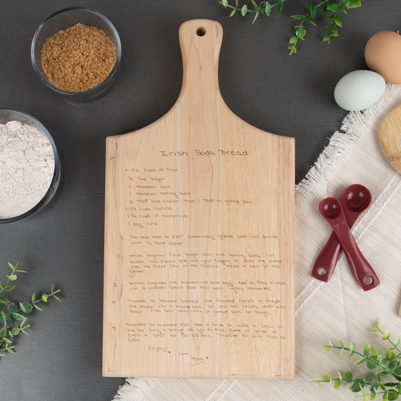 Maple paddle board shown engraved with a "Irish Soda Bread" recipe engraved on it. The handle of the paddle is at the top, & a few kitchen items such as flour, brown sugar, eggs, & measuring tools are displayed around the cutting board