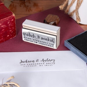 A white envelope flap stamped with a wooden hand stamp showing the return address in black ink. The wooden hand stamp lies on its side with the stamp plate inked next to a black ink pad.