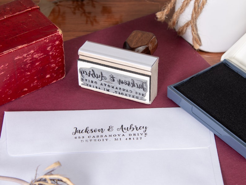 A white envelope flap stamped with a wooden hand stamp showing the return address in black ink. The wooden hand stamp lies on its side with the stamp plate inked next to a black ink pad.