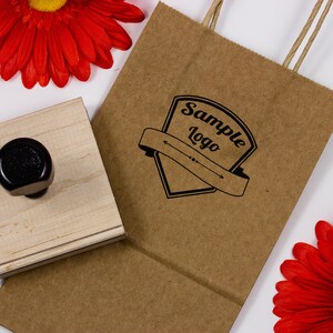 A sample logo is used as an impression in black ink onto a brown paper bag. A wood handle stamp sits next on top of the brown paper bag. Two red flowers are placed on both sides of the brown paper bag.