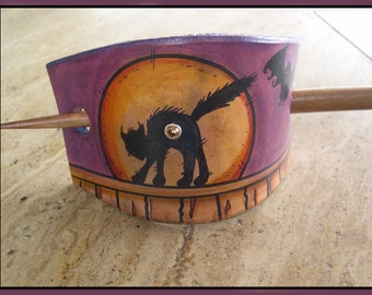Könnte beinhalten: Eine lila-orangefarbene Haarspange mit Halloween-Motiv. Die Spange zeigt eine schwarze Katzen-Silhouette vor einem Vollmond, mit einem Holzstab zur Befestigung der Haare. Die Spange ist etwa 10 cm lang.