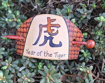 Könnte beinhalten: Eine handgefertigte Haarspange mit einem zentralen beigefarbenen Feld mit einem schwarzen und orangefarbenen Tigerzeichen und dem Text "Year of the Tiger". Die Spange hat einen braunen, strukturierten Rand und einen dekorativen Holzstab.