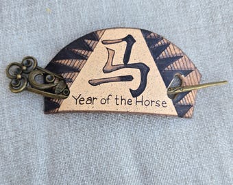 Könnte beinhalten: Haarspange aus Keramik mit beigem Hintergrund, schwarzen und braunen geometrischen Mustern und dem Text "Year of the Horse". Die Spange hat eine bronzefarbene dekorative Nadel.