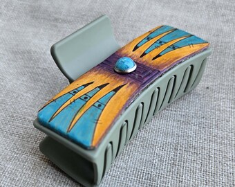 May include: A rectangular hair claw clip in shades of gray, with a decorative top. The top is painted with a design featuring blue and yellow shapes, a purple band, and a turquoise stone. The clip is approximately 3 inches long.