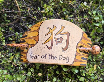Könnte beinhalten: Haarspange aus Keramik mit beigem Hintergrund und braunen und gelben Akzenten. Die Spange zeigt das chinesische Schriftzeichen für "Hund" und den Text "Year of the Dog". Ein Holzstab ist eingesetzt.