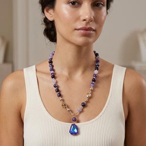May include: A beaded necklace featuring a variety of purple and lavender spherical beads, interspersed with small gold-toned metallic accents. The necklace includes a large, faceted, iridescent blue pendant suspended from a gold-toned metal clasp.