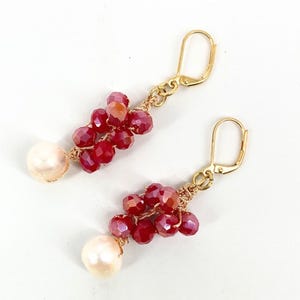 Peut inclure: Une paire de boucles d'oreilles pendantes avec des fermoirs dormeuses dorés, des grappes de perles en cristal facetté rouge foncé et une perle ronde blanc cassé à la base, le tout assemblé par un fin fil métallique de couleur dorée.