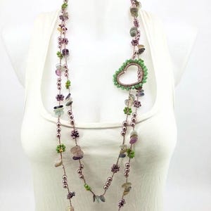 Può includere: Una lunga collana a più fili con perline color malva, piccole perline verdi a forma di fiore e accenti traslucidi simili a pietre irregolari. Un ciondolo a forma di cuore, bordato da piccole perline verdi, è appeso alla collana.