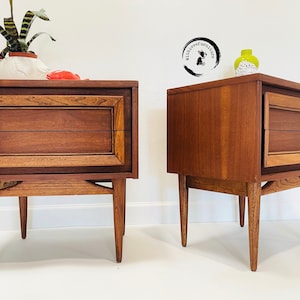 May include: Two matching mid-century modern wooden nightstands with two drawers each. The nightstands have a dark wood finish and are on four legs.