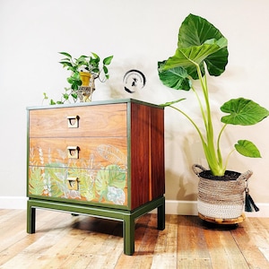 May include: A three-drawer dresser with a green painted base and a wood top. The drawers are decorated with a tropical leaf design. The dresser is in front of a white wall with a potted plant to the right.